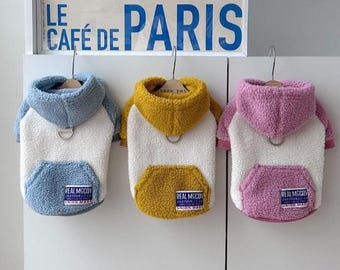 May include: Three dog hoodies in blue, yellow, and pink, each with a white body and a kangaroo pocket. The hoodies have a fluffy texture and a small tag that reads "REAL MCCOY". The background includes a sign that says "LE CAFÉ DE PARIS".