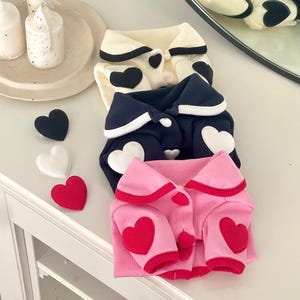 May include: Three pet sweaters in cream, navy, and pink, each with heart-shaped appliqués. The cream sweater has black hearts, the navy has white hearts, and the pink has red hearts. A black, white, and red heart-shaped patch are next to the sweaters.