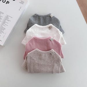 Może przedstawiać: Cztery koszulki z krótkim rękawem ułożone w stos, każda w innym neutralnym kolorze: szarym, białym, różowym i szarobrązowym. Każda koszulka ma okrągły dekolt i małą metkę z grafiką na ramieniu.