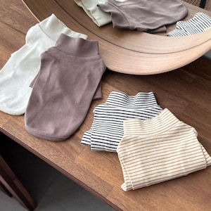 Könnte beinhalten: Auswahl an Rollkragenpullovern in verschiedenen Farben und Mustern. Enthält unifarbene Farben wie Weiß und Mauve sowie gestreifte Designs in Schwarz und Weiß sowie Beige und Weiß. Die Hemden sind ordentlich auf einer Holzoberfläche gefaltet.
