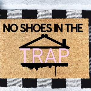 Peut inclure: Un paillasson en fibre de coco brun avec une bordure &agrave; carreaux noirs et blancs. Le texte "NO SHOES IN THE TRAP" est imprim&eacute; en noir sur le paillasson. Le mot "TRAP" est en rose et se trouve &agrave; l'int&eacute;rieur d'une silhouette noire d'une maison.