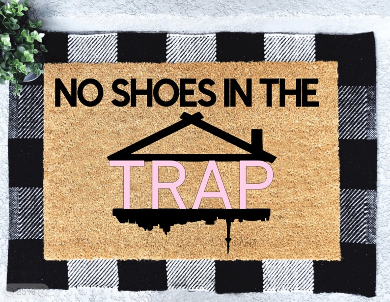 Peut inclure: Un paillasson en fibre de coco brun avec une bordure &agrave; carreaux noirs et blancs. Le texte "NO SHOES IN THE TRAP" est imprim&eacute; en noir sur le paillasson. Le mot "TRAP" est en rose et se trouve &agrave; l'int&eacute;rieur d'une silhouette noire d'une maison.