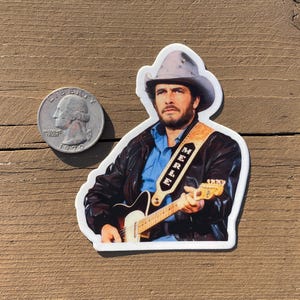 May include: A sticker featuring a man playing a guitar, wearing a cowboy hat and a leather jacket. The guitar strap reads "Merle". A U.S. quarter is next to the sticker for size comparison.