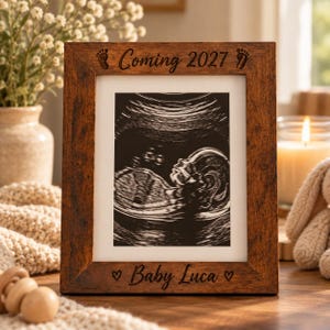 May include: A rectangular dark brown wooden picture frame featuring an ultrasound image of a fetus. The top of the frame is engraved with the text Coming 2027 and two baby footprints. The bottom is engraved with Baby Luca and two hearts.