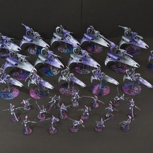 May include: A collection of purple and silver painted miniature spaceships and warriors. The spaceships are on circular bases with a purple and blue mottled design. The warriors are on smaller circular bases with a pink and purple mottled design. The miniatures are arranged in a formation on a gray surface.