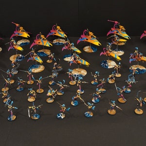 May include: A collection of miniature figures painted in vibrant colors, including pink, yellow, blue, and purple. The figures are arranged in a battle formation, with some figures on flying platforms. The figures are part of a tabletop wargame.