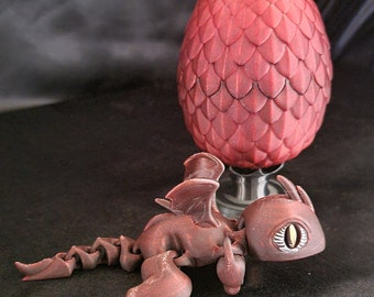 May include: A small, red dragon figurine with a segmented tail and wings, lying on its belly. The dragon has a large, yellow eye and is positioned in front of a red dragon egg.