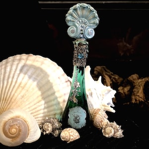 May include: A decorative green glass bottle with a seashell-shaped stopper, adorned with silver accents and a blue gem. The bottle is labeled "Mermaid Tears" and is surrounded by various seashells, creating a coastal theme.
