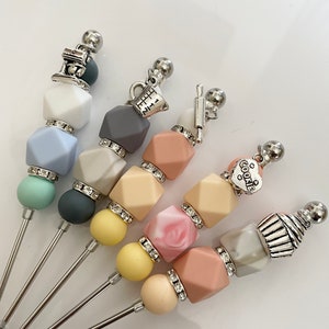 May include: Five colorful silicone and metal stirring sticks with various baking-themed charms. The charms include a mixer, measuring cup, rolling pin, heart with the word "cookie", and a cupcake.
