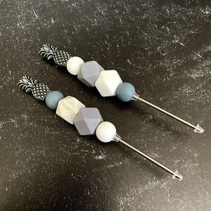 Pode incluir: Dois colares de contas de silicone cinza e branco com pingentes de abacaxi prateados e pontas de metal prateadas. Os colares estão sobre um fundo preto.