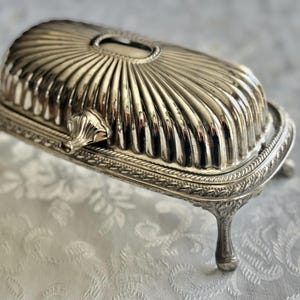 May include: Silver-colored butter dish with a ribbed, domed lid and ornate detailing. The dish has a rectangular base with decorative trim and rests on four curved feet. A small handle is on the side.