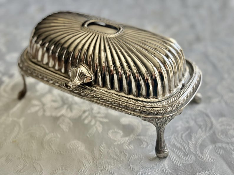 May include: Silver-colored butter dish with a ribbed, domed lid and ornate detailing. The dish has a rectangular base with decorative trim and rests on four curved feet. A small handle is on the side.