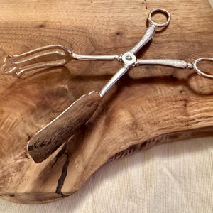 May include: Silver serving tongs with a decorative design on the handles. The tongs have a fork on one end and a spoon on the other.