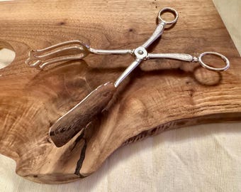 May include: Silver serving tongs with a decorative design on the handles. The tongs have a fork on one end and a spoon on the other.