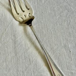 May include: A silver serving fork with four tines and a decorative handle. The fork has a detailed design on the head and a smooth, elongated handle. The fork is likely made of stainless steel or silver.