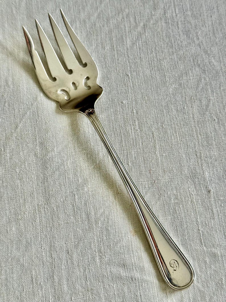 May include: A silver serving fork with four tines and a decorative handle. The fork has a detailed design on the head and a smooth, elongated handle. The fork is likely made of stainless steel or silver.
