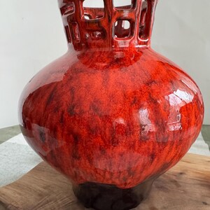 Puede incluir: Un jarr&oacute;n de cer&aacute;mica rojo brillante con un cuerpo bulboso y una base marr&oacute;n oscuro. La parte superior presenta un dise&ntilde;o geom&eacute;trico calado. El jarr&oacute;n se exhibe sobre una superficie de madera, mostrando su color vibrante y forma &uacute;nica.