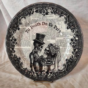 K&ouml;nnte beinhalten: Schwarzer und wei&szlig;er Keramikteller mit einem gotischen Hochzeitsthema. Der Teller zeigt zwei Skelette in Hochzeitskleidung mit dem Text "Til Death Do Us Part" in einer dekorativen Schrift.