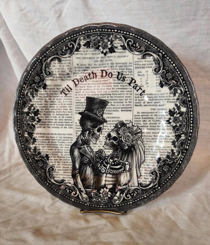 K&ouml;nnte beinhalten: Schwarzer und wei&szlig;er Keramikteller mit einem gotischen Hochzeitsthema. Der Teller zeigt zwei Skelette in Hochzeitskleidung mit dem Text "Til Death Do Us Part" in einer dekorativen Schrift.