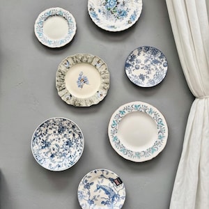 Op de afbeelding: Een verzameling van zeven keramische borden gerangschikt aan een muur, met diverse blauw-witte bloemen-, botanische en vogelpatronen. De borden vari&euml;ren in grootte en randdetails, waaronder geschulpte en gladde randen met gedetailleerde ontwerpen.