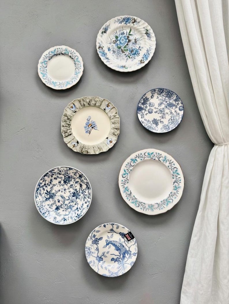Op de afbeelding: Een verzameling van zeven keramische borden gerangschikt aan een muur, met diverse blauw-witte bloemen-, botanische en vogelpatronen. De borden vari&euml;ren in grootte en randdetails, waaronder geschulpte en gladde randen met gedetailleerde ontwerpen.
