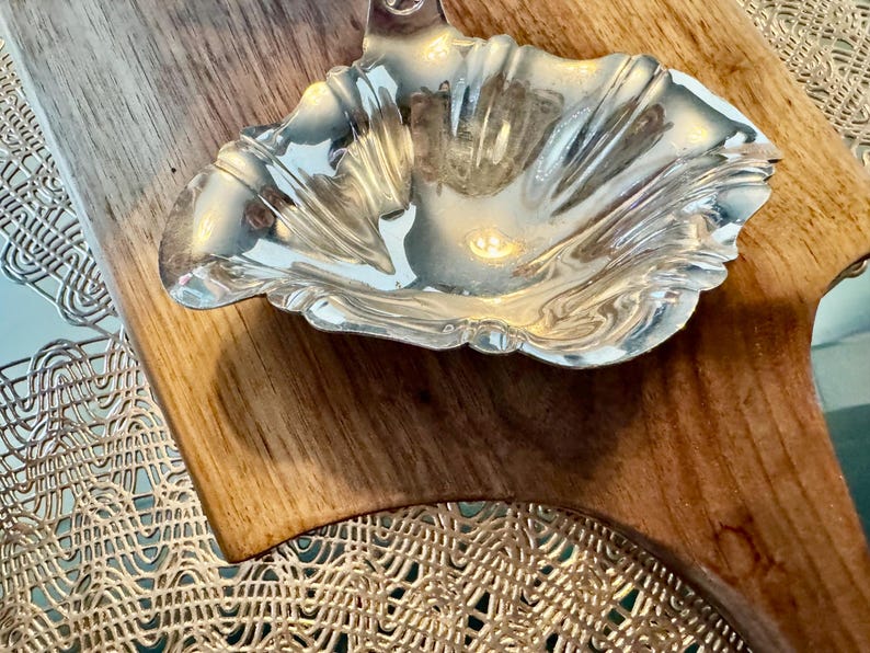 May include: A silver, ornate, decorative dish with a scalloped edge sits on a wooden surface.