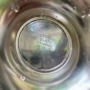 May include: Close-up of a silver-colored metal object with the text "VICTORIAN PLATE E.P. COPPER 229" etched into the center. The object has a circular design with water droplets on the surface. The item appears to be vintage.