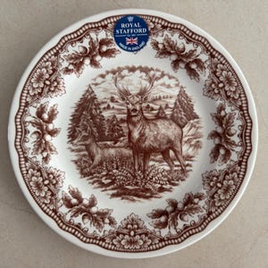 Op de afbeelding: Een wit keramieken bord met een bruine transferprint van een hert en een hinde in een bosrijke omgeving. De rand is versierd met een herhalend patroon van eikenbladeren en eikels. Een blauwe sticker vermeldt Royal Stafford Est 1845.
