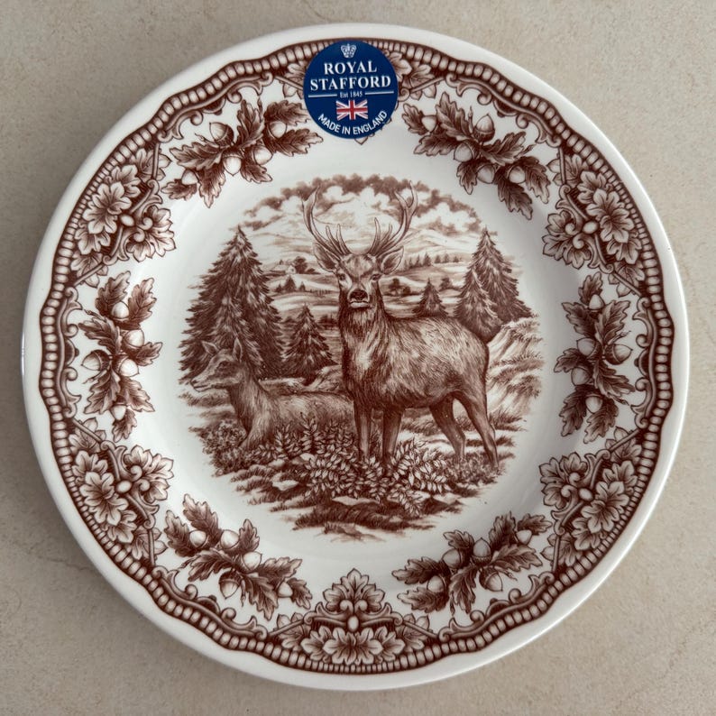 Op de afbeelding: Een wit keramieken bord met een bruine transferprint van een hert en een hinde in een bosrijke omgeving. De rand is versierd met een herhalend patroon van eikenbladeren en eikels. Een blauwe sticker vermeldt Royal Stafford Est 1845.