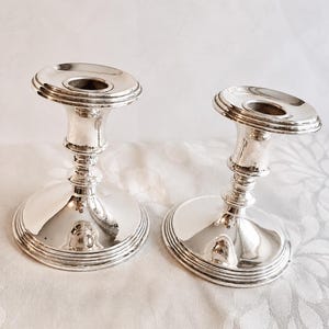 Pu&ograve; includere: Una coppia di portacandele in metallo color argento lucido caratterizzati da un'ampia base circolare con anelli concentrici a gradini, uno stelo centrale sottile e un bordo superiore svasato per sostenere una candela affusolata.
