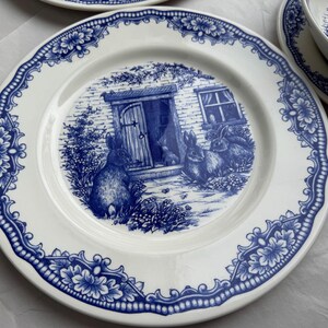K&ouml;nnte beinhalten: Ein wei&szlig;er Keramikteller mit einem blauen und wei&szlig;en floralen Rand und einer Szene von Kaninchen vor einem H&auml;uschen. Die Szene ist in Blau und Wei&szlig; bemalt.
