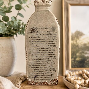 May include: A beige ceramic vase with a distressed, crackled finish and a rectangular shape. The vase features handwritten script and decorative accents in brown. The vase is displayed on a wooden surface, with a framed picture and a plant in the background.