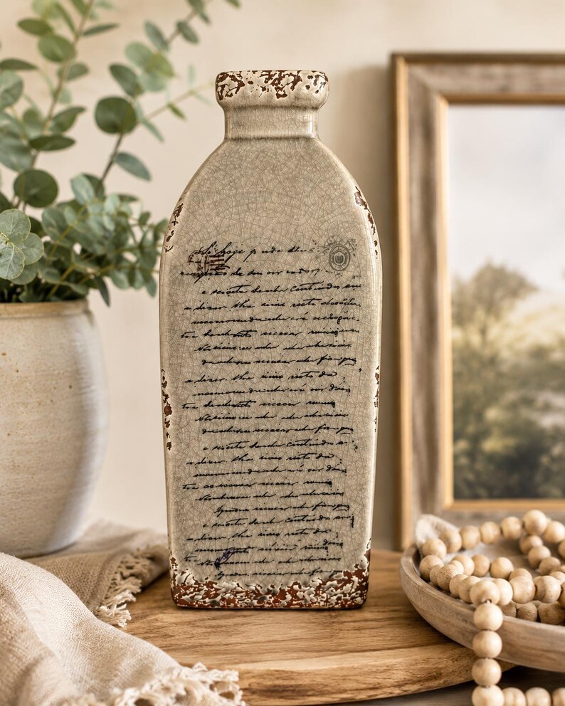 May include: A beige ceramic vase with a distressed, crackled finish and a rectangular shape. The vase features handwritten script and decorative accents in brown. The vase is displayed on a wooden surface, with a framed picture and a plant in the background.