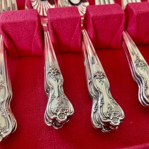 May include: A set of silver-toned metal flatware arranged in a storage case with a vibrant red velvet interior. Each handle features an ornate, embossed floral pattern at the base, with smooth, curved stems leading up to the utensil heads.