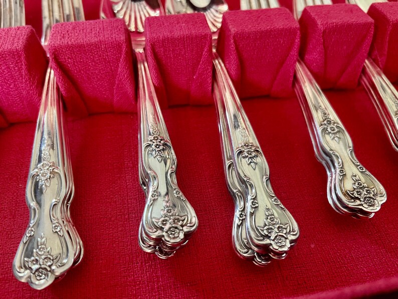 May include: A set of silver-toned metal flatware arranged in a storage case with a vibrant red velvet interior. Each handle features an ornate, embossed floral pattern at the base, with smooth, curved stems leading up to the utensil heads.