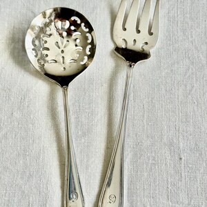 May include: A silver serving spoon with a decorative pierced bowl and a silver fork. The spoon has a long handle and a floral design in the bowl. The fork has four tines. Both utensils have a small emblem at the base of the handle.