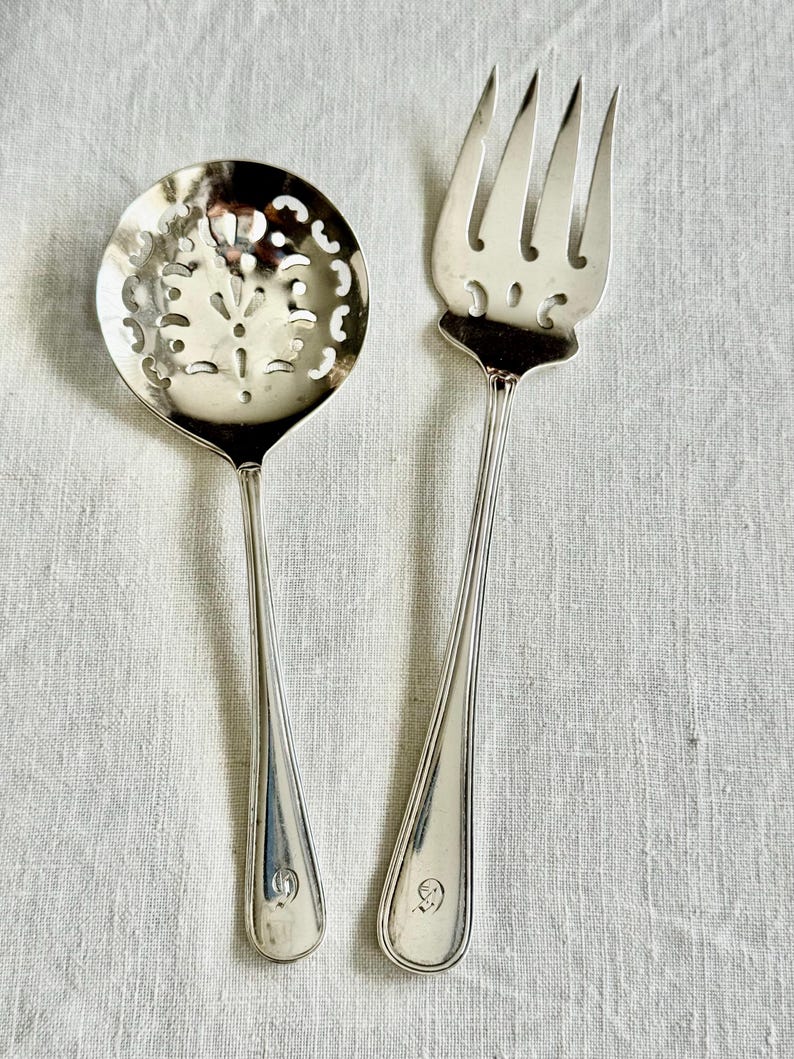 May include: A silver serving spoon with a decorative pierced bowl and a silver fork. The spoon has a long handle and a floral design in the bowl. The fork has four tines. Both utensils have a small emblem at the base of the handle.