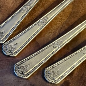 Pu&ograve; includere: Primo piano di quattro cucchiai antichi color argento. Ogni cucchiaio presenta un design ornato sul manico, che include un elemento a forma di cuore e dettagli floreali. I cucchiai sono disposti diagonalmente, mostrando i loro intricati motivi.
