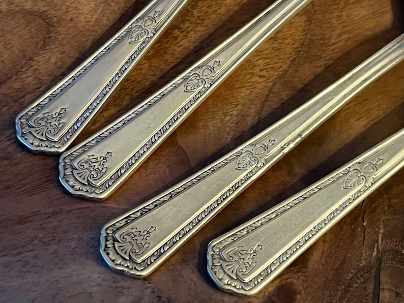 Pu&ograve; includere: Primo piano di quattro cucchiai antichi color argento. Ogni cucchiaio presenta un design ornato sul manico, che include un elemento a forma di cuore e dettagli floreali. I cucchiai sono disposti diagonalmente, mostrando i loro intricati motivi.