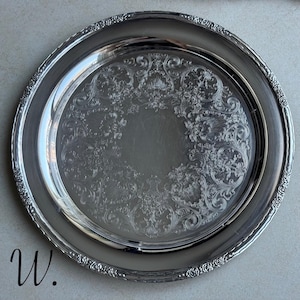 May include: A round silver-colored metal serving tray featuring an intricate embossed floral and scrollwork pattern around the center and a decorative textured border along the rim. The letter W is printed in black in the bottom left corner.