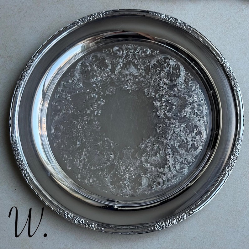 May include: A round silver-colored metal serving tray featuring an intricate embossed floral and scrollwork pattern around the center and a decorative textured border along the rim. The letter W is printed in black in the bottom left corner.