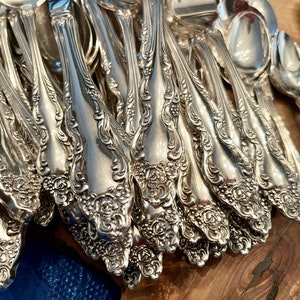 May include: A collection of ornate silver-colored silverware, including forks, spoons, and knives. The silverware features a detailed floral pattern on the handles. The items are arranged on a wooden surface, showcasing the intricate designs and reflective qualities of the metal.