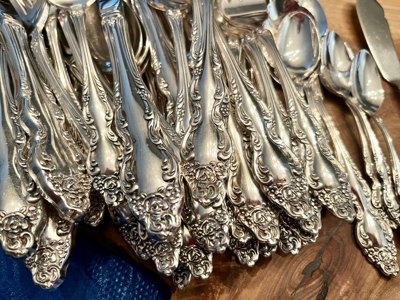 May include: A collection of ornate silver-colored silverware, including forks, spoons, and knives. The silverware features a detailed floral pattern on the handles. The items are arranged on a wooden surface, showcasing the intricate designs and reflective qualities of the metal.