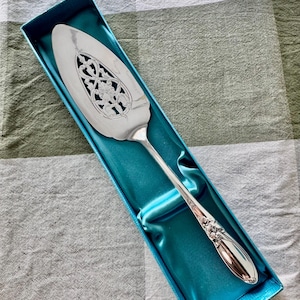 Op de afbeelding: Zilveren taartschep met een sierlijk, opengewerkt ontwerp in het midden, gepresenteerd in een turquoise blauwe doos. De handgreep heeft bloemendetails. De schep is ongeveer 25 cm lang.