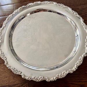 May include: A round silver-toned serving tray featuring a polished, reflective center surface and an ornate, scalloped border decorated with embossed floral and scrollwork patterns along the entire outer rim of the metal platter.