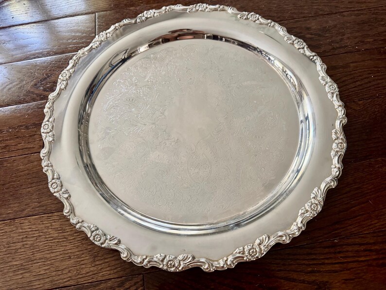 May include: A round silver-toned serving tray featuring a polished, reflective center surface and an ornate, scalloped border decorated with embossed floral and scrollwork patterns along the entire outer rim of the metal platter.