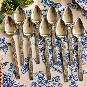 May include: A set of eight silver-toned grapefruit spoons with serrated tips and rectangular handles featuring an embossed geometric pattern. The spoons are arranged in a vertical formation, showcasing their polished metal finish and detailed decorative stems.