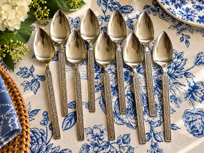 May include: A set of eight silver-toned grapefruit spoons with serrated tips and rectangular handles featuring an embossed geometric pattern. The spoons are arranged in a vertical formation, showcasing their polished metal finish and detailed decorative stems.
