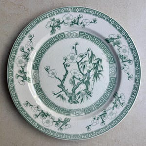 May include: A round ceramic plate with a cream-colored base and a fine crackle glaze finish. The plate features a dark green transfer-printed design of flowering plum branches and bamboo, with a geometric border pattern around the center.