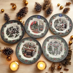 May include: A collection of decorative plates with Christmas scenes. Each plate features a different winter holiday image, surrounded by a floral border. The plates are arranged with pine cones, gold ornaments, and lit candles.